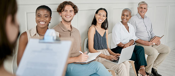 Five people are sitting in a row, facing a person holding a clipboard. They are smiling and holding notebooks or a laptop, suggesting they are in a meeting or interview setting. The background is neutral with light-colored walls.