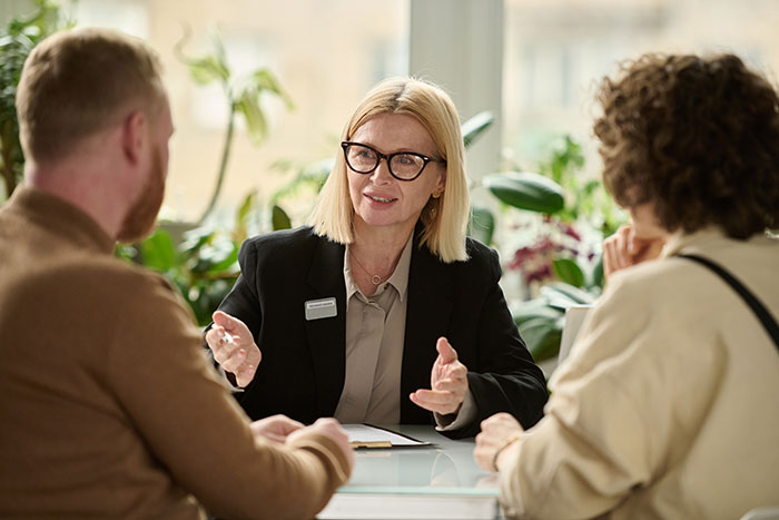 A woman with glasses and a name tag gestures while talking to two people at a table. She is in a business setting with plants in the background. The people are engaged in conversation.