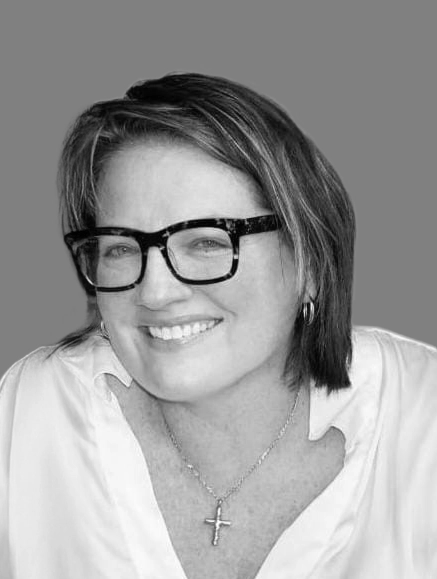 A woman with short, dark hair wears large, dark-framed glasses, a light-colored shirt, and a cross necklace. She is smiling and the background is a solid gray.