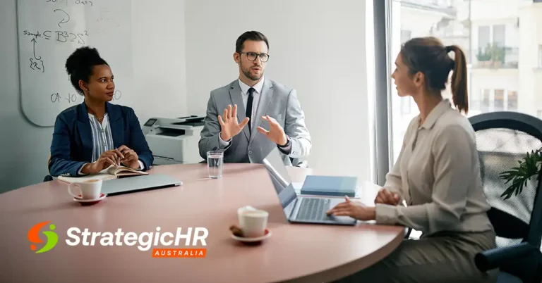 Three professionals are seated at a round table in an office, having a discussion. One person is speaking with hands raised, while another takes notes and the third uses a laptop. The StrategicHR Australia logo is visible.