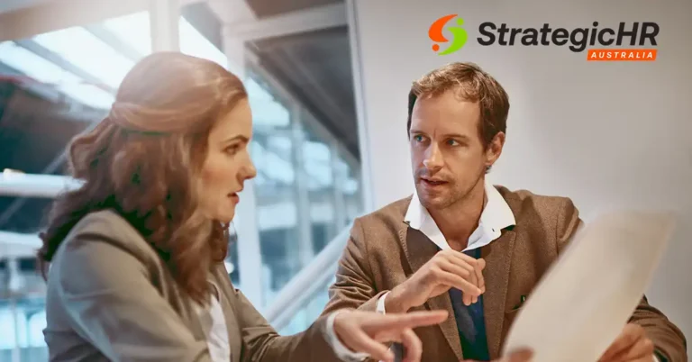 A man and a woman sit at a desk in an office, engaged in a discussion about the performance management process while looking at a document. The StrategicHR Australia logo appears in the top right corner of the image.