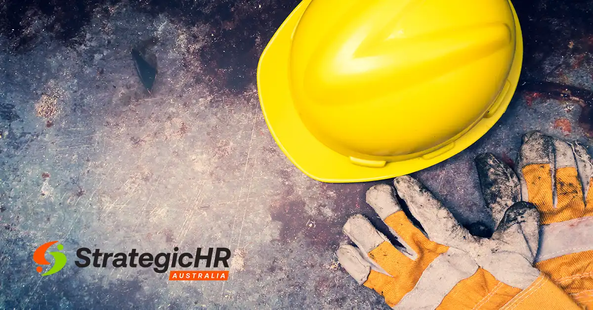 A yellow safety helmet and a pair of orange work gloves rest on a scratched metal surface. The StrategicHR Australia logo appears in the lower left corner.