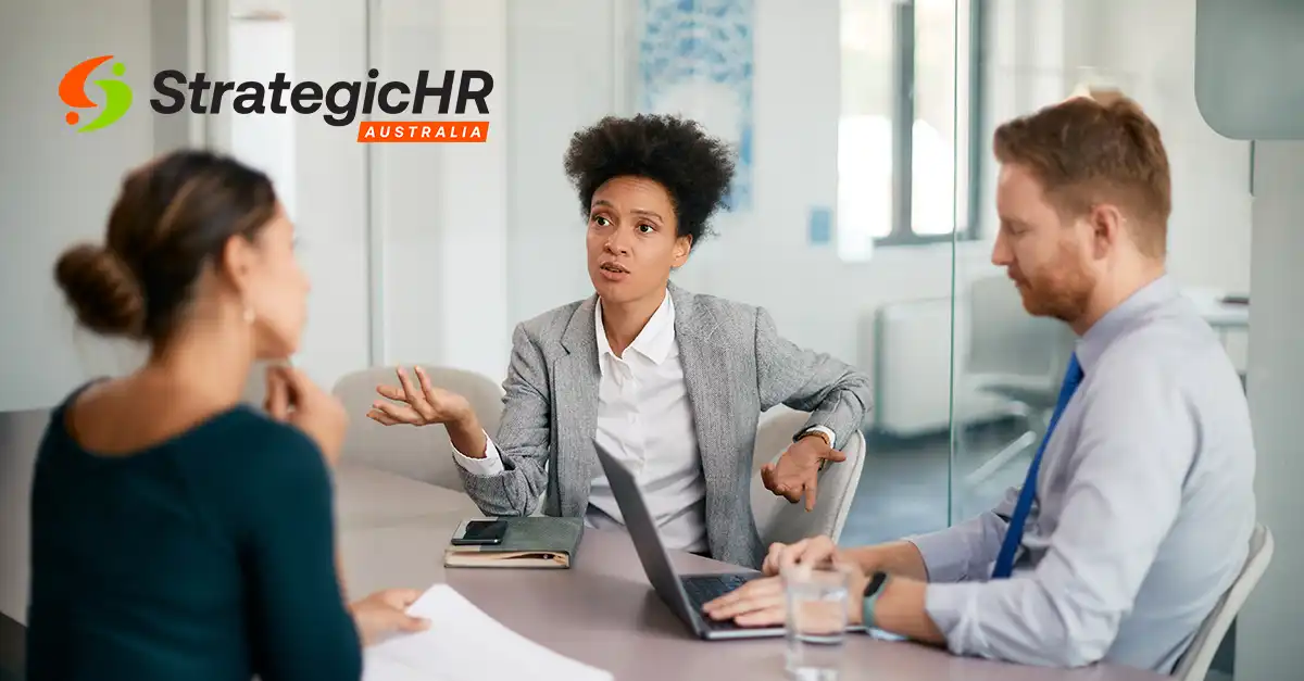 Three people are seated at a table in a modern office, engaged in a discussion. One woman gestures with her hand while speaking. The "StrategicHR Australia" logo is visible in the top-left corner.