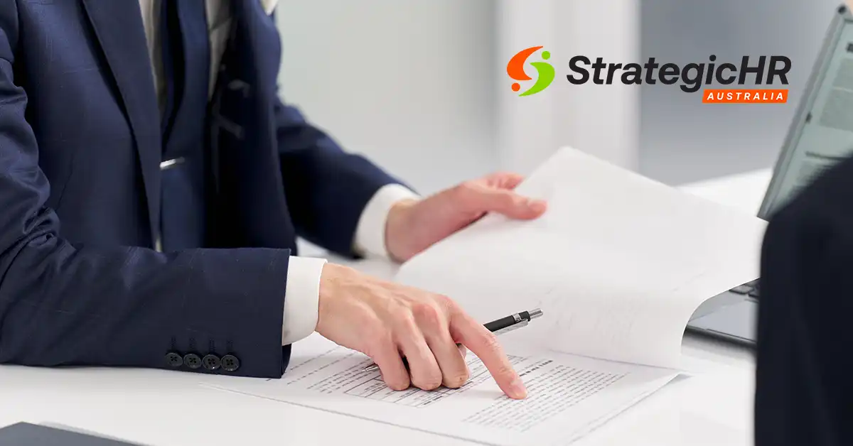 A person in a suit points to a document on a desk, holding a pen, with a laptop nearby. The StrategicHR Australia logo is visible in the upper right corner of the image.