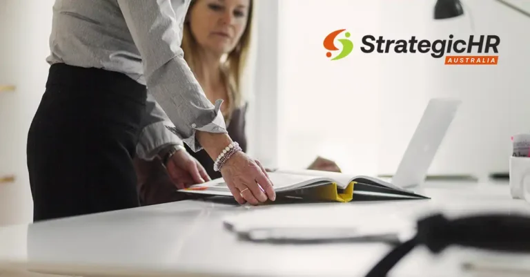 Two people work at a desk with an open binder and a laptop. One person points at the binder. The StrategicHR Australia logo is visible on a wall in the background.