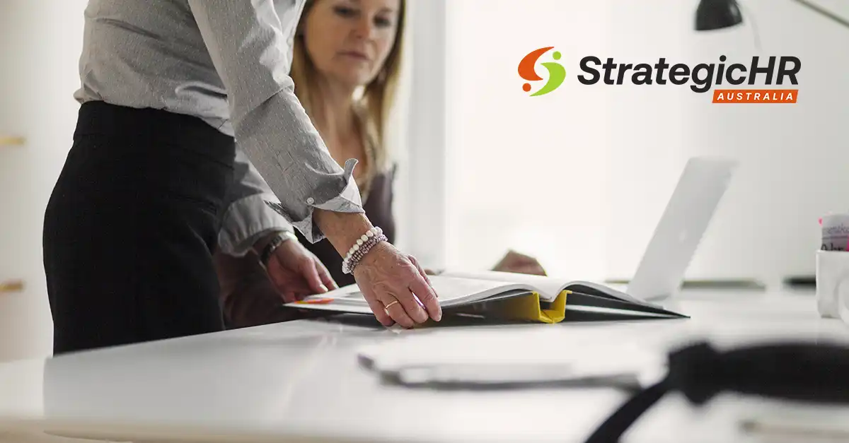 Two people work at a desk with an open binder and a laptop. One person points at the binder. The StrategicHR Australia logo is visible on a wall in the background.