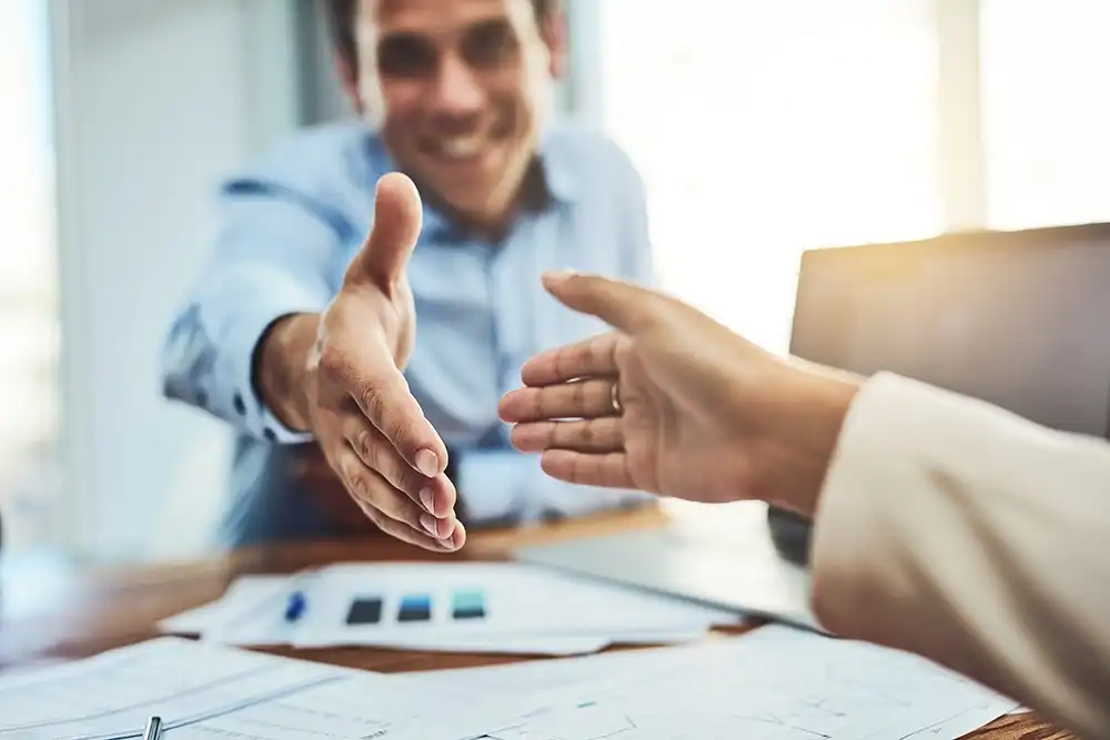 Two people are reaching out for a handshake across a desk with papers and a laptop, suggesting a business meeting or agreement.