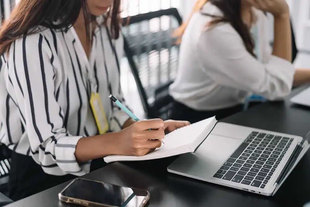Two women sit at a desk; one writes in a notebook with a pen while a laptop, smartphone, and other office items are on the table. Both are wearing business attire.