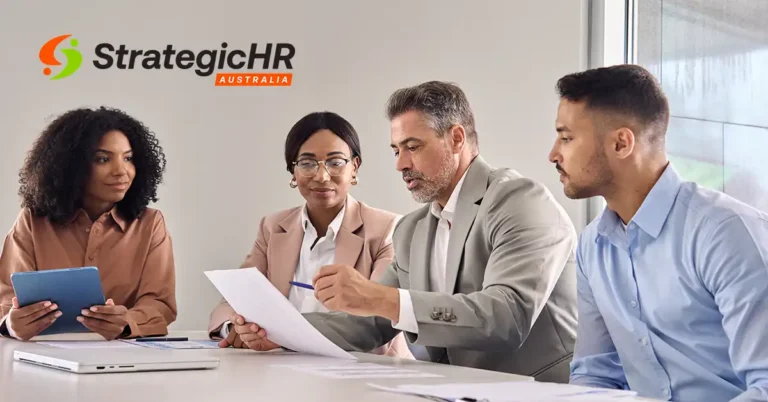 Four professionals sit at a table in a meeting room, reviewing documents together. One man points to a paper, while another man and two women listen. The Strategic HR Australia logo is visible on the wall.