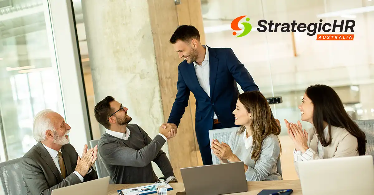 Four people in business attire sit at a conference table with laptops, two men shake hands, and two women applaud. The "StrategicHR Australia" logo is visible on a glass wall in the background.