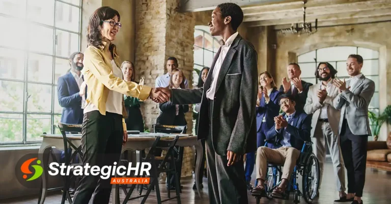 Two people shake hands in an office setting while colleagues applaud in the background. Some people are seated, including one person in a wheelchair. The StrategicHR Australia logo is visible in the lower left corner.