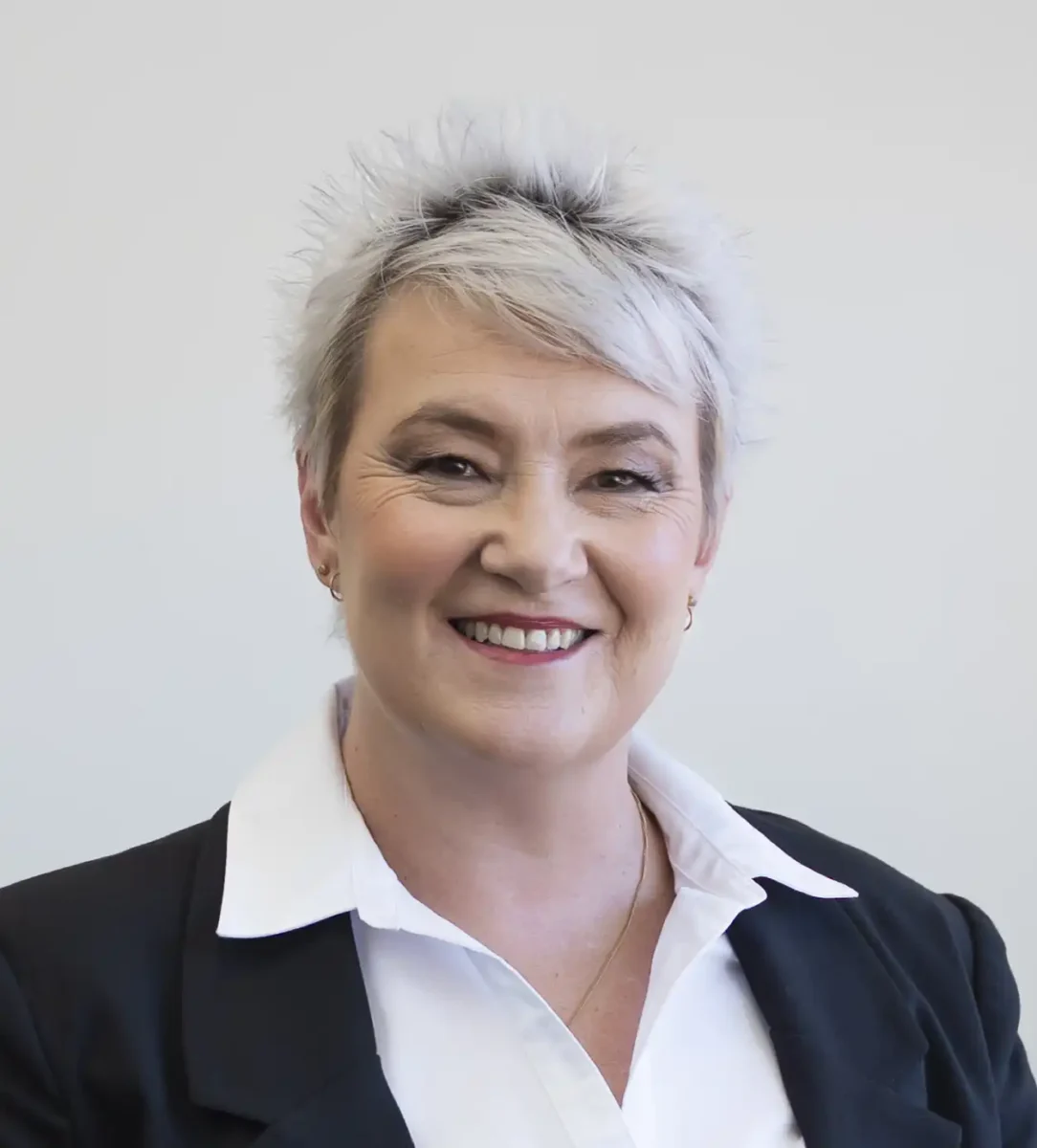 A person with short, spiky gray hair is wearing a white collared shirt and a black blazer, smiling in front of a plain light-colored background.