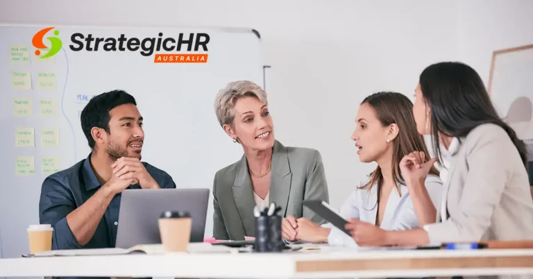 Four people sit at a table having a discussion with laptops and coffee cups. A whiteboard with sticky notes and the StrategicHR Australia logo are visible in the background.
