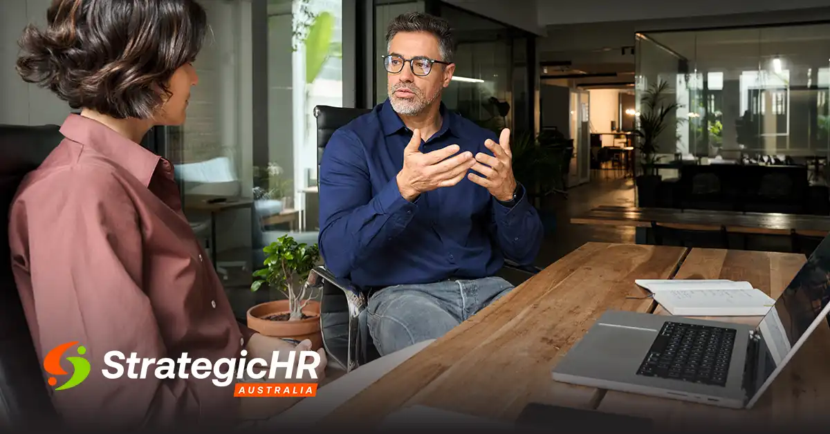 Two people sit across from each other at a wooden table in a modern office, having a discussion. An open laptop and documents are on the table. The StrategicHR Australia logo is visible in the foreground.