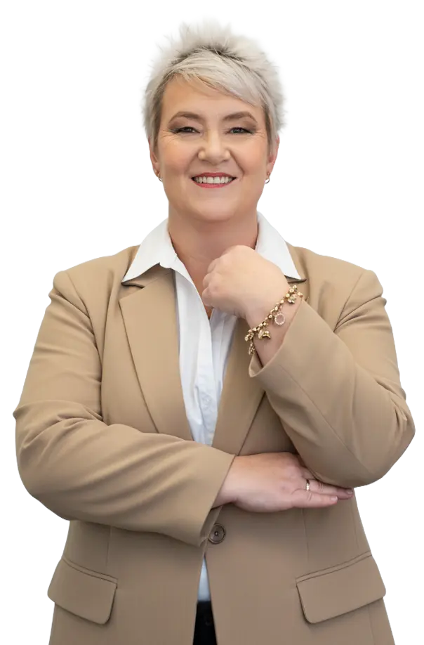 A person with short, light gray hair is standing and smiling, wearing a tan blazer over a white collared shirt. One arm is crossed while the other hand, with a bracelet, is raised to chest level.