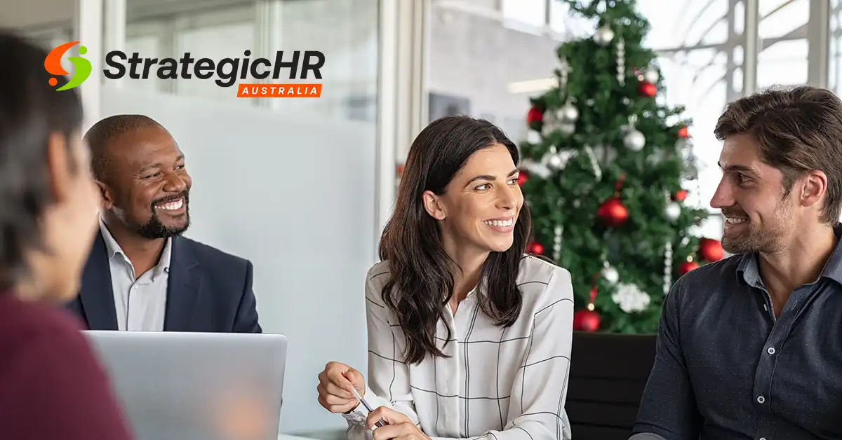Four people sit around a table in an office, engaged in conversation about employee wellbeing. A decorated Christmas tree is in the background. The Strategic HR Australia logo is visible in the top left corner.