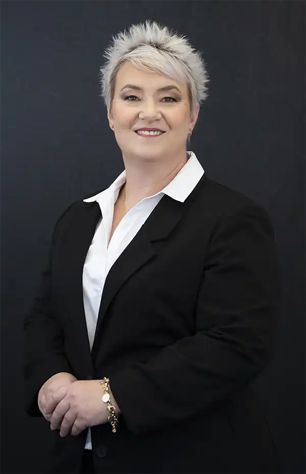 A person with short, spiky, gray hair wears a black blazer over a white collared shirt, standing against a plain dark background and smiling at the camera.