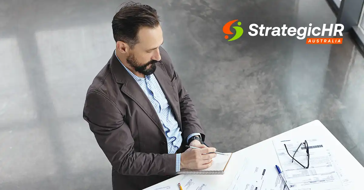A man in a brown blazer sits at a desk with papers, a notebook, and glasses. He is holding a pen and looking down. The StrategicHR Australia logo is displayed in the top right corner.
