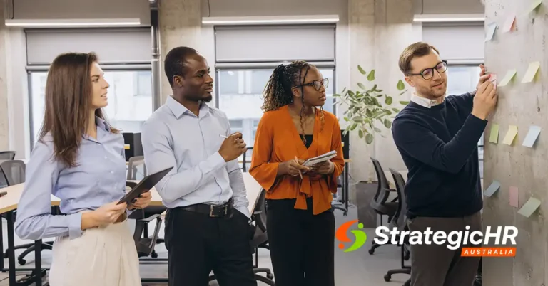 Four people in business attire stand in an office, with one man placing sticky notes on a wall while the others observe and take notes. The Strategic HR Australia logo is visible in the bottom right corner.