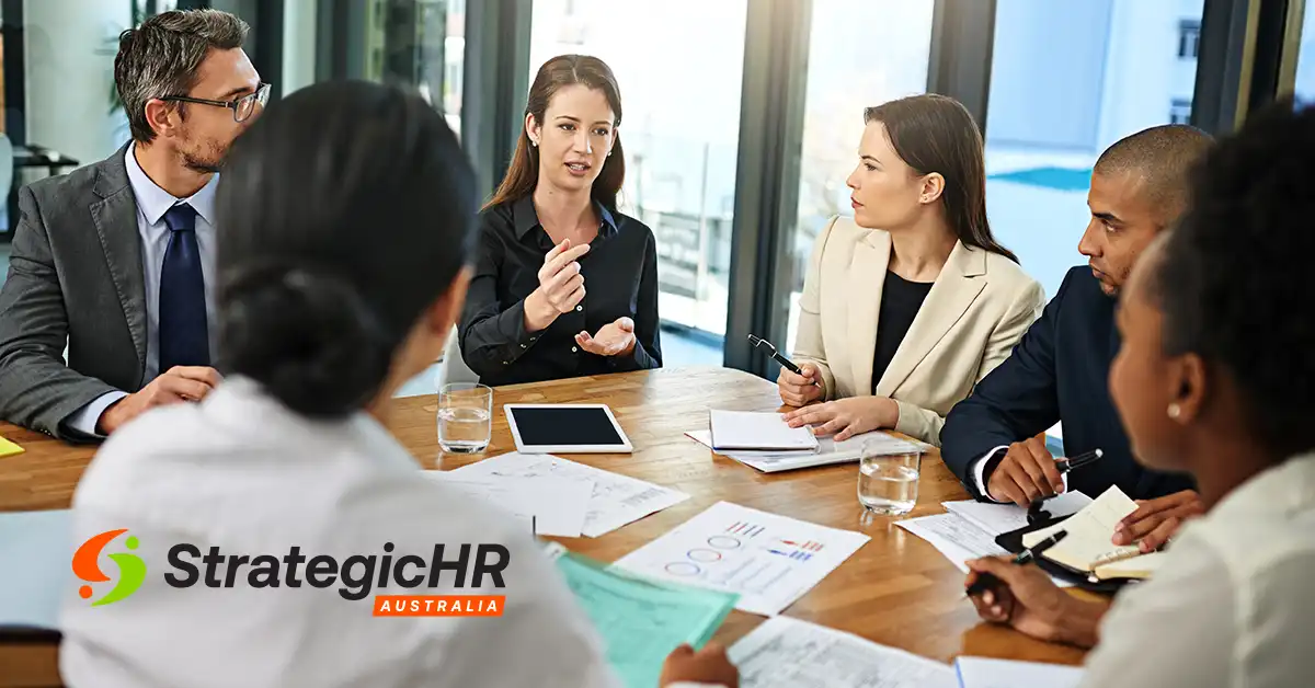 A group of six professionals sit around a table in an office, discussing documents and charts. A tablet is on the table. The Strategic HR Australia logo is visible in the lower left corner, highlighting HR clarity for leaders.