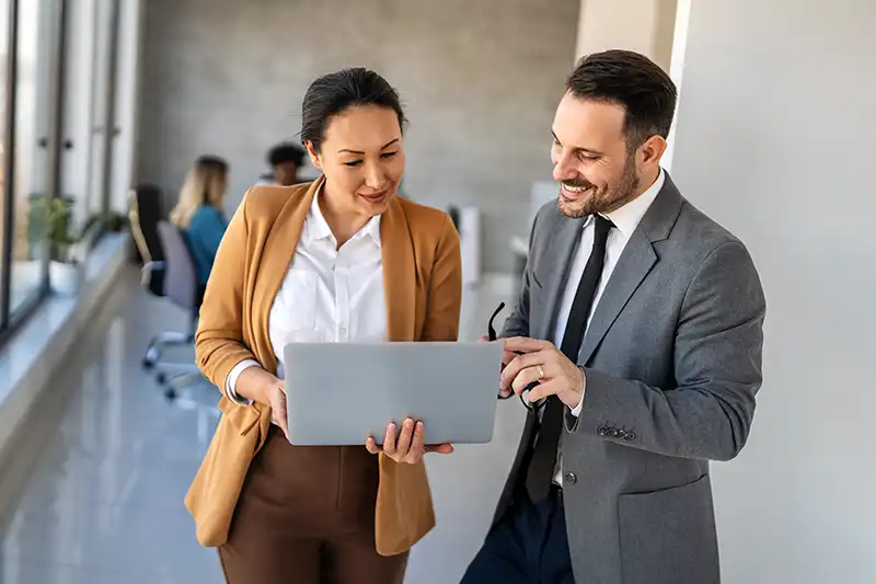 Two business professionals stand in an office hallway, looking at a laptop and discussing something while smiling. Other people are working at desks in the background.