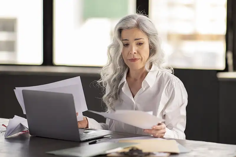 A woman with long gray hair wearing a white shirt sits at a desk, looking at papers in her hands. An open laptop and more documents are on the desk in front of her. She appears focused.