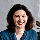 A woman with shoulder-length brown hair is smiling. She is wearing a blue top and is seated in front of a light gray textured wall with a leafy plant partially visible on the side.