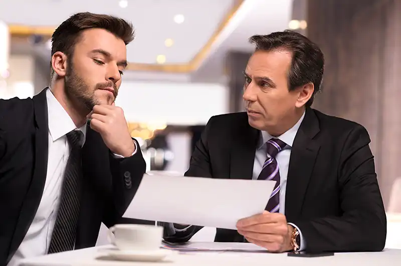 Two men in business suits sit at a table indoors, reviewing a document. One is explaining details related to workplace investigations while the other listens thoughtfully with his hand on his chin.