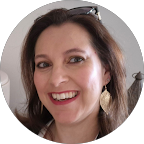 A woman with long brown hair smiles at the camera. She is wearing gold leaf-shaped earrings, red lipstick, and sunglasses resting on her head. The background includes a lamp and a gray wall.