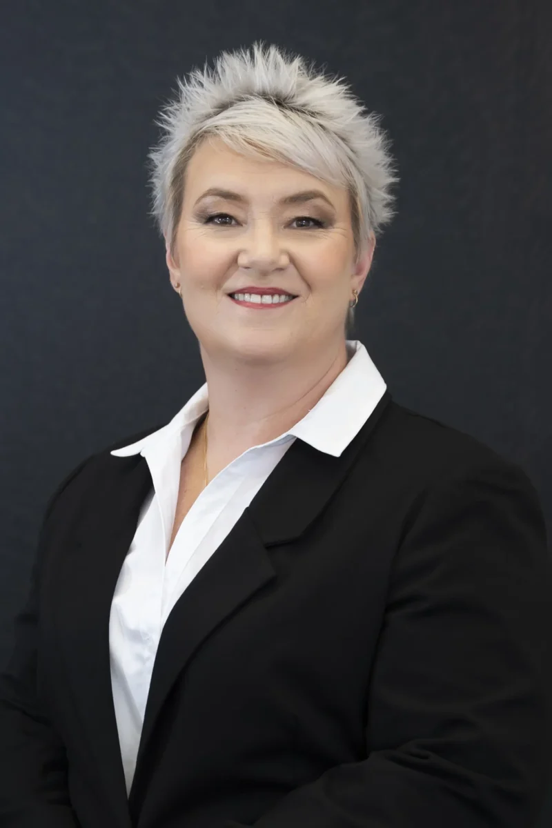 A person with short, spiky gray hair is wearing a black blazer over a white collared shirt and is smiling at the camera against a plain dark background.