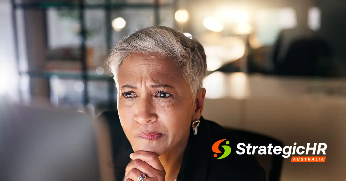 A person with short gray hair looks thoughtfully at a computer screen, reflecting on psychosocial risk obligations in Australia. The Strategic HR Australia logo is in the bottom right corner. The background is an office setting with blurred lights.