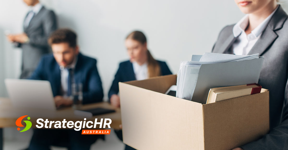 A person in business attire holds a box of office supplies and documents, while two colleagues sit at a table with laptops in the background. The Strategic HR Australia logo is visible on the lower left.