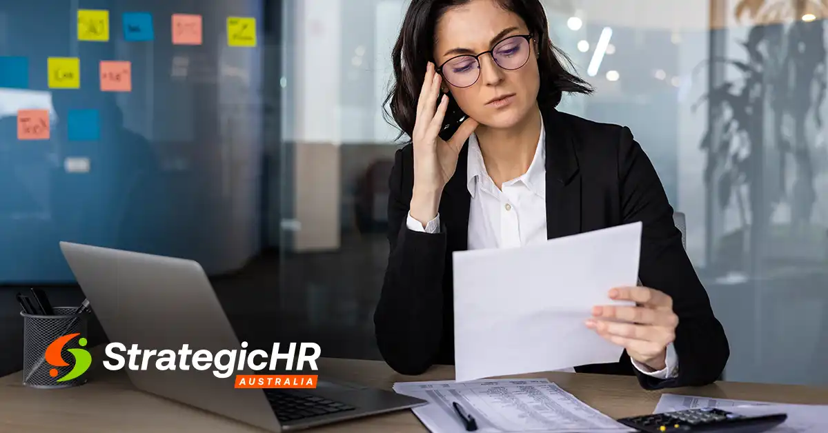 A woman in business attire sits at a desk with a laptop, calculator, and documents, holding a paper and talking on the phone. The StrategicHR Australia logo appears in the lower left corner.