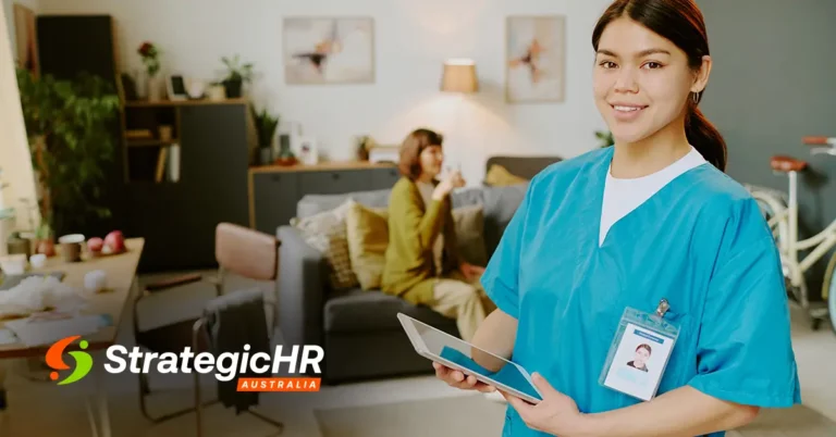 A healthcare worker in scrubs holds a tablet and smiles at the camera in a living room, reflecting on SCHADS Award sleepover shifts. In the background, another woman sits on a couch. The Strategic HR Australia logo appears in the lower left corner.