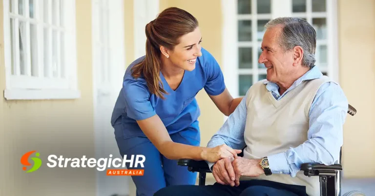 A female caregiver in blue scrubs smiles and holds hands with an older man sitting in a wheelchair. The StrategicHR Australia logo is visible in the lower-left corner. They are outside near a building with windows.