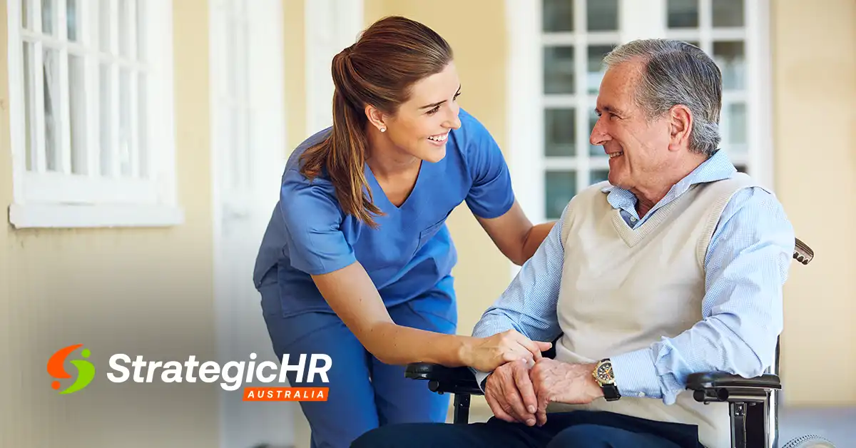 A female caregiver in blue scrubs smiles and holds hands with an older man sitting in a wheelchair. The StrategicHR Australia logo is visible in the lower-left corner. They are outside near a building with windows.
