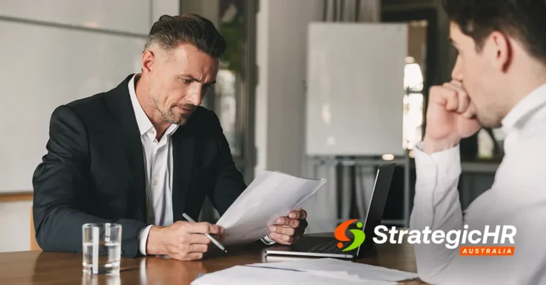 Two men in business attire sit at a table in an office, discussing documents and psychosocial risk in HR processes. One reads papers as the other listens. The StrategicHR Australia logo is in the corner.