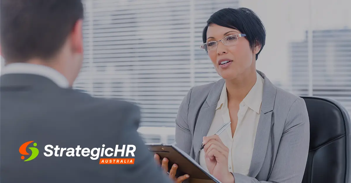 A woman in business attire holds a clipboard and pen while talking to a man in an office. The StrategicHR Australia logo is visible in the lower left corner.
